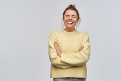 Portrait of cheerful, adult girl with blond hair gathered in bun. Wearing yellow sweater. Laughing with eyes closed and arms crossed on a chest. Stand isolated over white background