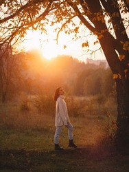 A young woman in a warm sweater stands under a large tree in an autumn park. Sunny autumn sunset