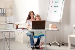 Mother sitting with her daughter on her lap at a table in a home classroom teaching her on a laptop computer