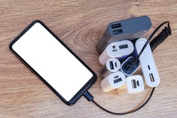 Set of power banks with a telephone on a wooden table. A portable charger charges a smartphone with a white screen mockup