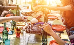 Group of happy friends eating and drinking beers at barbecue dinner on sunset time - Adult people having meal together outdoor - Focus on fork sausages - Summer lifestyle, food and friendship concept