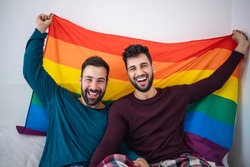 Gay couple holding lgbt rainbow flag indoors on bed at home - Focus on right man face