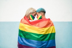 Young couple of lesbian women under lgbt rainbow flag wearing masks at gay pride parade - Focus on faces