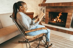 African woman reading book in front of cozy fireplace at home - Winter lifestyle concept - Focus on face