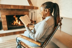 African woman reading book in front of cozy fireplace at home - Autumn and winter lifestyle concept - Focus on ear