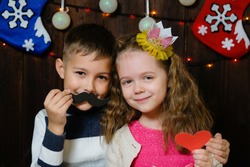 Kids with props for a photo booth. Little boy and girl with the requisite mustache, heart and crown on christmas wooden background. Event, holiday, party. Children congratulate happy new year.