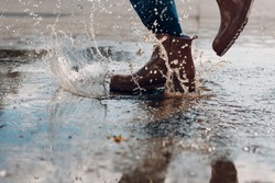 Woman wearing rain rubber boots walking running and jumping into puddle with water splash and drops in autumn rain.