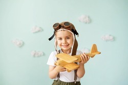 A boy in an aviator helmet and suspenders stands on a blue background with clouds and plays with a wooden plane.