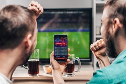 Two male buddies watching live football game broadcast on tv and celebrating money win after making bets online at bookmakers website.