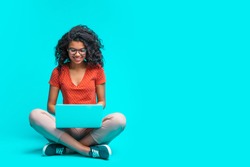 Beautiful young smiling woman in casual outfit and trendy eyeglasses sitting isolated on bright colored blue background and working on her laptop. Full length horizontal studio shot. Copy space.