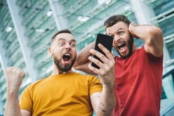 Two friends showing sincere emotions of joy about victory in online lottery. Men being happy winning a bet in online sport gambling application with football stadium on the background.