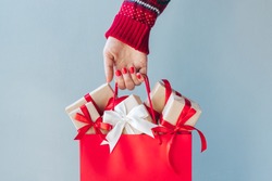 Cropped image of female hand with red polished nails holding shopping bag full of christmas gift boxes. Holiday sale concept. 