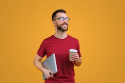 Portrait of confident young smiling bearded man in casual red t-shity and trendy spectacles holding laptop and paper cup of coffee, isolated on yellow background.