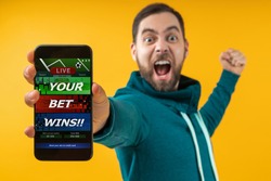 Man being happy winning bet in online sport gambling application on mobile phone, screaming and clencing his his fist. Close up cropped shot. Focus is on hand. Isolated over yellow background.