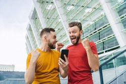 Two bearded guys showing celebrating victory in online lottery. Friends being happy winning a bet in online sport gambling application with football stadium on the background.
