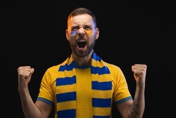 Portrait of euphoric happy soccer supporter man in yellow-blue t-shirt and scarf and painted face, cheering for his favourite team, isolated over black background.