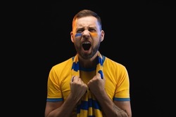 Portrait of excited young soccer supporter man in yellow-blue t-shirt and scarf and painted face, cheering for his favourite team. Isolated over black background.