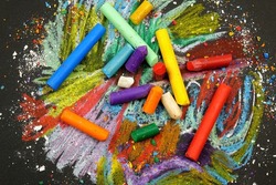 Multi-colored wax crayons lie on a black surface with chaotic crayon drawings on it