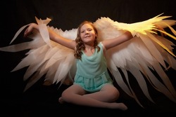 A young girl in a green dress and with white wings is dancing in a dark studio with a bright flash. A model posing for a photo shoot like an angel