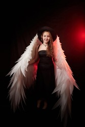 A young elegant teenage girl in black dress and with white wings in a dark studio with bright flash. A model posing for a photo shoot like an angel