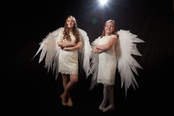 Two teenage girl in white dress and with white wings in a dark studio with bright flash. Sisters and models posing for a photo shoot like an angel