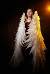 A young elegant teenage girl in black dress and with white wings in a dark studio with bright flash. A model posing for a photo shoot like an angel