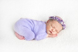 Newborn. Newborn baby. A baby girl is sleeping on a light bed. A newborn baby girl in a wreath and a light woolen suit of purple is sleeping on a light-colored blanket. Pose of the baby.