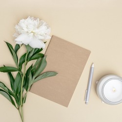 Feminine desktop, closed notebook, pen, fresh white peony flower and lit fragrant candle in glass jar on beige background with copy space. Summer work concept.