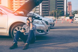 Young asian handsome man sitting by the Broken Down car and using smart phone for help