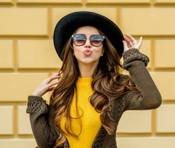 Fashion woman in a hat and sunglasses, in knit dress and jacket outdoors in the fall.Blowing lips kiss.