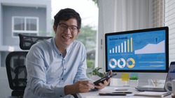 Portrait head shot of young attractive asian man sitting smiling work multiple screen computer and smart tablet on table desk at home in concept freelance data analyst, data scientist for business.