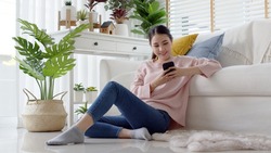Young attractive beautiful casual asia female lady student look at cellphone play social media sit at sofa couch living room feel relax comfort carefree with green houseplant at cozy home apartment.