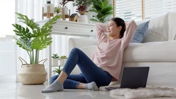 Young attractive beautiful asia female girl or university student sit smile look outside window put arm hand back behind head at sofa couch living room feeling relax comfort at cozy home houseplant.