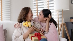 Attractive beautiful asian middle age mum sit with grown up daughter give gift box and flower in family moment celebrate mother day. Overjoy bonding cheerful kid embrace relationship with retired mom.