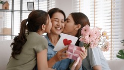 Mother day two grown up child cuddle hug give flower gift box red heart card to mature mum. Love kiss care mom asia middle age adult three people sitting at home sofa happy smile enjoy family time.