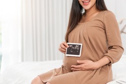 Asian pregnant woman in brown dress holding ultrasound on her belly in bed at bedroom. Pregnancy, parenthood, preparation and expectation concept