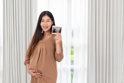 Asian pregnant woman in brown dress holding ultrasound on her belly in bed at bedroom. Pregnancy, parenthood, preparation and expectation concept