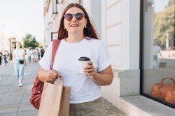 Beautiful fashionable young woman in sunglasses with shopping paper bag standing on city street, black friday. Urban lifestyle concept. Attractive female drinking a coffee in paper bio white cup.