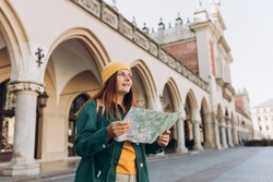 Attractive young female tourist is exploring new city. Redhead girl in hat holding a paper map on Market Square in Krakow. Traveling Europe in autumn. High quality photo