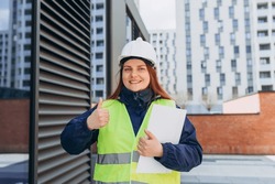 Happy Architect redhead woman wearing contractor white helmet and safety yellow vest holding project paper plan over building background doing ok sign, thumb up with finger, Modern exterior