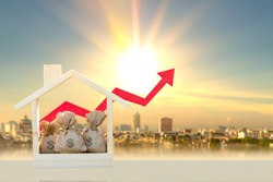 Money bags in the wooden home model and red arrow graph with growing value on photo blur cityscape background, Loan and business investment for real estate concept.
