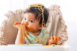 Adorable little child funny girl eating spaghetti with spoon while sitting in high-powered chair at home. Toddler child with tomato sauce making mess her face looking at parent. Self-feeding concept