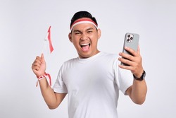 Excited young Asian men celebrate Indonesian independence day on 17 August while holding the Indonesian flag and mobile phone isolated over white background