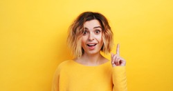 Portrait happy young woman slow motion of finding solution Idea pointing forward an index finger on isolated yellow background, looking directly at the camera smiling. Monotone. Emotions of people