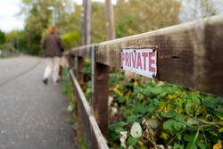 Very shallow focus of a Private sign attached to a wooden fence, leading to a private river mooring area. A woman and her dog are in the distance, going for a walk.