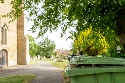 Shallow focus of a green wheelie bin used for dead flowers in a rural English cemetery. A church entrance can be seen on the left.