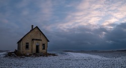 Outside of a dilapidated yellow-brick one-room school house located on the open Canadian prairie.  Picture taken at dusk with white and blue-grey clouds and a hint of pink. Snow on the field.