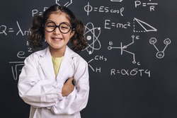smiling little girl science student with glasses in lab coat on school blackboard background with hand drawings science formula pattern, back to school and successful female career concept
