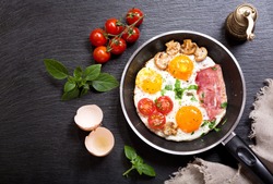 breakfast with fried eggs in a pan with bacon and vegetables on dark background, top view