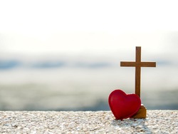 Red heart with wooden Christian cross on gravel floor in morning light, beach sea as background. Jesus love you. Faith hope believe in God. Believe in salvation. Christianity background concept.